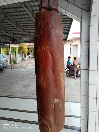 The image shows a large, wooden, cylindrical object with a dark brown hue. It is hanging from the ceiling of a covered structure. The object appears to be an old, weathered piece of wood. Carved into its side are the numbers "2012," indicating it was likely made or installed that year. The background is a blurry image of a building in Kecamatan Gabuswetan, Indonesia. Two people are visible in the background, a man in a blue shirt and a woman in a pink shirt. The man is sitting on a motorcycle, and the woman is sitting behind him. The motorcycle is parked in a courtyard. The ground is paved with white tiles.
