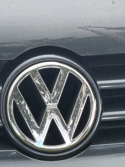 A close-up, low-angle, slightly blurry photograph shows the front of a dark grey Volkswagen car. The focus is on the chrome Volkswagen logo, which is circular and has a silver "VW" emblem in the center. The logo is mounted on the car's grille, which consists of horizontal black slats. The car's hood is visible in the upper portion of the image, reflecting a light grey sky and some faint horizontal lines. The image appears to have been taken outdoors on a cloudy day in Bemmel, Netherlands.