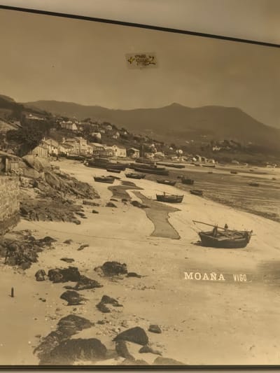 A vintage, sepia-toned photograph captures a tranquil coastal scene in Moaña, Spain. The image depicts a wide sandy beach at what appears to be low tide, revealing extensive stretches of wet sand and numerous dark rocks scattered across the foreground and along the shoreline. Many small fishing boats are beached on the sand, some partially submerged or resting near a winding tidal channel that crosses the beach.

Along the left side, a stone wall and rocky terrain lead up to a hillside where a village is nestled. Numerous buildings, characteristic of a coastal town, line the shore and ascend the hillside, with more structures visible further up. Distant mountains form a hazy backdrop to the scene. The sky is clear, suggesting calm weather conditions, and no people are visible in the photograph.

In the bottom right corner of the image, the text "MOAÑA VIGO" is clearly visible in white block letters, identifying the location as Moaña, likely in relation to the nearby city of Vigo. A small, partially obscured sticker with illegible text is present in the upper center of the photograph.