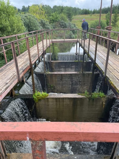 An overhead view shows a multi-tiered dam on a cloudy day. A wooden walkway with rusted red railings extends over the dam, with a person fishing at the far end of the walkway. Water cascades down the concrete tiers of the dam, creating whitewater rapids at the bottom. Lush green trees and foliage surround the dam and walkway. The scene appears to be located in Bereznik, Russia.