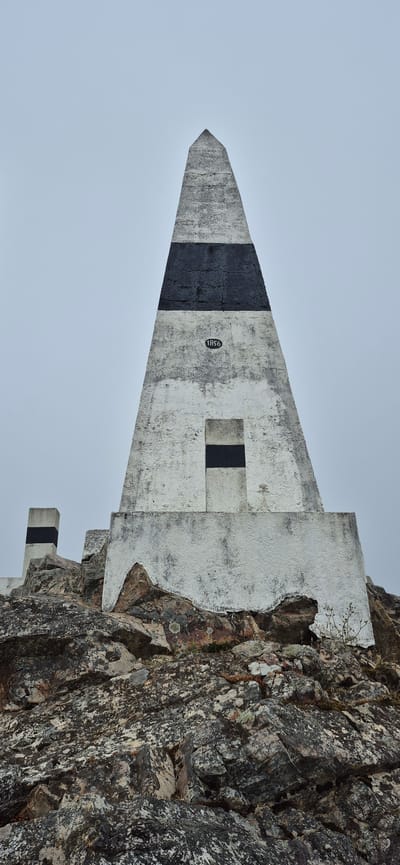 The image shows a concrete obelisk with black and white bands, situated on a rocky outcrop under an overcast sky. The obelisk is roughly triangular in shape and tapers to a point. There is a rectangular indentation with a black band on its face, and the number "1856" is inscribed in a circle to the left of this indentation. A smaller, similar concrete marker with black and white bands is visible to the left of the obelisk, also on the rocky terrain. The rocks are varied in color and texture, with patches of lichen and sparse vegetation. The scene appears to be a landmark or monument in an outdoor, elevated location. This monument is located at Monte da Madalena in Póvoa de Varzim, Portugal, and it marks the geographical location of the city.