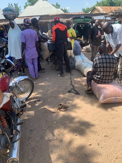 This media file depicts a scene in Boi A, Nigeria, with several individuals engaged in the loading or unloading of large sacks, likely containing goods, onto or from a vehicle. The overall setting appears to be an outdoor market or roadside area, characterized by a dirt ground, some rudimentary structures with corrugated metal roofs, and vegetation in the background.

The subjects are a group of men, some of whom are actively handling the sacks. One man wears a plaid shirt and is partially obscured by a large sack. Another man in a black t-shirt with a graphic on it appears to be directing or observing the activity. Several other men are visible in various states of participation, including one wearing a bright yellow shirt and another in a purple tunic. The presence of motorcycles in the foreground and background suggests transportation is a key element of the scene, likely used to bring the goods to this location or to transport them away.

The time of day appears to be daytime, as indicated by the strong shadows cast on the ground, suggesting bright sunlight. The weather seems to be warm and dry, consistent with an outdoor market environment in Nigeria. The mood seems to be one of industrious activity, with people focused on their tasks.

One of the sacks has some visible text on it, including what appears to be "SACKS" in large letters. Other markings on the sacks are less legible. The overall impression is of a bustling, working environment where goods are being moved, possibly for trade or distribution.