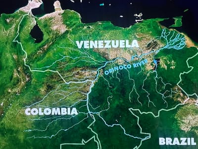 This media file is a satellite image showing a portion of South America. The image displays a vast, green landscape dominated by intricate river systems, highlighted in light blue. The Orinoco River is prominently labeled, flowing through Venezuela and appearing to originate or connect with tributaries in Colombia and Brazil, as indicated by the text labels for these countries. The terrain shows varied topography, with some mountainous or elevated areas appearing in lighter shades of green and brown. The image is primarily focused on geographical features and political boundaries, with no people or specific activities depicted. The overall impression is one of a lush, expansive, and geographically significant region, emphasizing its extensive waterways.