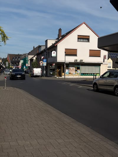 This image depicts a street scene in Sankt Augustin, Germany, likely during the daytime given the clear blue sky with wispy clouds and the bright sunlight casting shadows. The perspective is from street level, looking along a road lined with buildings.
The primary subjects are buildings and vehicles. On the left side of the road, a multi-story building with a white facade and a red-tiled roof dominates the foreground. It features several windows with brown shutters. Below this, a row of smaller buildings, also with pitched roofs, is visible. One of these buildings has a green awning and signage indicating it's an "APOTHEKE" (pharmacy). Another building further down the street also has a similar sign.
Several vehicles are parked along the street or driving. A silver car is parked near the pharmacy, and a white van is further down. A black car with German license plates ("SU-MY 280") is driving on the road. In the background, more buildings and vehicles can be seen, suggesting a small urban or suburban environment.
Notable details include various street signs, such as parking signs with the letter "P" and other informational symbols. A bird is flying in the sky on the left side of the image. The road surface appears to be asphalt, and the sidewalk on the right is paved with cobblestones. The overall impression is one of a typical day in a German town. There is no discernible activity or event taking place beyond normal street life.