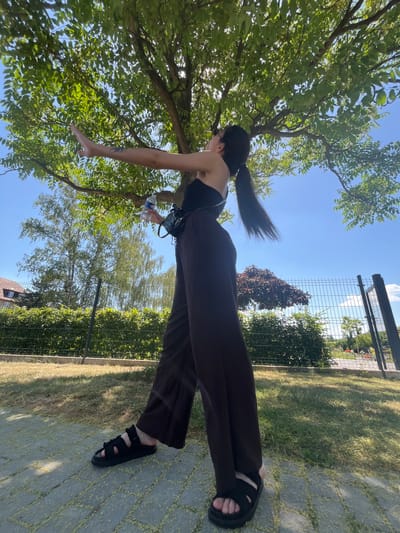 A person with dark hair in a ponytail is shown in a side-view, low-angle shot outdoors. They are wearing a black strapless top and wide-legged dark trousers, with black sandals on their feet. The person appears to be in motion, with one arm extended outwards. They are holding a clear plastic bottle of water in their hand.

The setting is sunny, with a clear blue sky overhead. Lush green trees with dense foliage are prominent, with sunlight filtering through the leaves. In the background, a chain-link fence separates the area from a grassy lawn and a paved walkway made of rectangular stones. Some bushes and a red-leafed tree are also visible behind the fence. The overall impression is a bright, warm day in a park-like or garden setting. The low angle of the shot emphasizes the sky and the trees, and the person's movement.