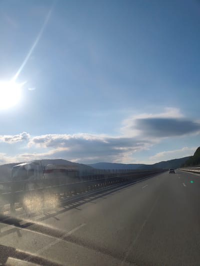 A sunlit highway stretches into the distance under a bright blue sky with scattered clouds. On the left, two large trucks, possibly a transport truck and a tanker truck, are parked or stopped on the side of the road. A highway guardrail runs along the shoulder, separating the road from the vehicles. In the background, rolling hills covered in vegetation fade into the horizon. The road itself is paved and marked with white lines. A single dark-colored car is visible ahead on the highway, traveling in the same direction. The scene appears to be a snapshot of travel on a highway in Bulgaria, specifically on the A2 Hemus highway near Vrachesh. The sun's glare is prominent, especially in the upper left quadrant, indicating a clear day with strong sunlight. The overall impression is one of a typical highway scene during daylight hours.