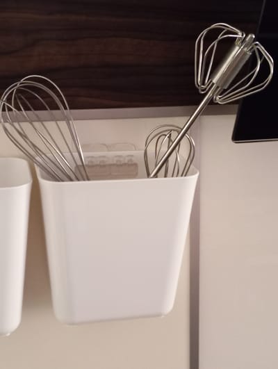 The image shows a close-up of two metal whisks, likely for a hand mixer, stored in a white container. The container is cylindrical and open at the top. One whisk is almost entirely contained within the holder, with only its handle and some of the wire loops visible. The second whisk is positioned above the container, with its wire loops extending outward, showcasing its intricate design. The handle of this upper whisk is metallic and appears to have some cylindrical attachments near its base.
The background of the image is divided into two distinct areas. To the left, a dark, vertically paneled surface suggests wooden cabinetry or a dark wooden door. To the right, a lighter, smooth surface, likely a wall or cabinet door, is visible. A thin horizontal stripe, possibly a metallic trim or a seam, runs across this lighter surface. The lighting appears to be artificial and even, with no strong shadows, suggesting an indoor setting.
No people are visible in the image, and the activity is implied to be storage of kitchen utensils. The setting appears to be a kitchen or a utility area. There is no visible text in the image. The overall impression is one of neat organization of kitchen tools. The location context of Baden, Austria, does not provide specific visual cues within the image itself.