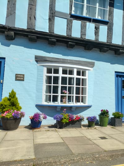 The image shows the exterior of Corbel House, a building located in Ludlow, United Kingdom. The house is painted a light blue and features traditional timber framing with dark wooden beams. A large white-framed window with multiple panes is visible in the center of the facade. A small rectangular sign with "CORBEL HOUSE" written on it is mounted on the wall to the left of the window. Several potted plants with colorful flowers are arranged on the paved sidewalk in front of the house. The sky is clear and the scene appears to be illuminated by sunlight.