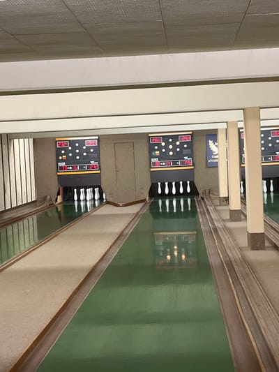 The image displays an unoccupied indoor nine-pin bowling alley (Kegelbahn) in Tuttlingen, Germany, featuring three lanes viewed from the approach area towards the pins. The ceiling is tiled in light beige, supported by horizontal beams and vertical square pillars of a similar light cream color. The background wall is a plain light brown/beige.

The three bowling lanes are constructed with a light beige/grey approach area transitioning into a long, reflective dark green surface leading to the pin deck. Dark wooden borders delineate the edges of the lanes and approach areas.

Above the end of the left and middle lanes are electronic scoreboards. The left scoreboard shows "272" and "310" at the top, and "73", "61", "19", "272" at the bottom, with a German flag icon and illuminated yellow and red indicators alongside a representation of pin positions. The middle scoreboard displays "146" and "293" at the top, and "38", "41", "1", "146" at the bottom, also with a German flag icon, indicators, and pin representations. A third scoreboard is partially visible on the right.

White bowling pins are set up on each visible lane. The left lane shows some pins dispersed, suggesting a recent ball throw, while the middle and right lanes have a full set of nine pins arranged in a diamond formation.

Between the left and middle lanes, a closed, light brown door is set into the back wall. Between the middle and right lanes, a blue sign with gold lettering including "Goll" and "»Somm", along with a gold wreath emblem, is mounted on the wall. The highly reflective green lane surfaces display clear inverted reflections of the pins, scoreboards, and overhead lighting fixtures. No individuals or bowling balls are present in the scene.