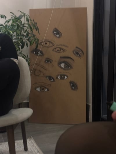 A rectangular artwork displays numerous eyes drawn in varying sizes and orientations against a tan background. The artwork is positioned near a plant and a chair. Part of a person’s body is partially obscuring the artwork. A string runs diagonally across the upper left of the artwork. The artwork appears to be framed or mounted, with a visible black frame at the top. The lighting in the scene is dim.