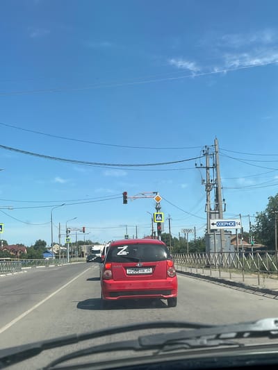This is a photograph taken from inside a vehicle, looking forward through the windshield. The perspective suggests the vehicle is in motion on a paved road in Ikryanoye, Russia. The sky is a clear, bright blue with some wispy clouds. Overhead, multiple power lines are strung between utility poles. In the middle ground, traffic lights are visible, one showing red and another potentially green in the distance. A red Kia Picanto car is in the foreground, in the same lane as the photographer's vehicle. The license plate of the red car reads "H086HK 30". A stylized "Z" is visible on the rear window of the red car. A pedestrian crossing sign with a yellow triangle and a walking person icon is present on the left side of the road. Further down the road, a sign with a black arrow pointing left and the Cyrillic text "ОЗЕРНОЕ 14" is visible, indicating a destination and distance. A "СТОП" (STOP) sign is also visible on the right side of the road. Buildings with red roofs and residential architecture are seen on the left side of the road. A metal railing runs along the side of the road. Trees and vegetation are present in the background on the right.
