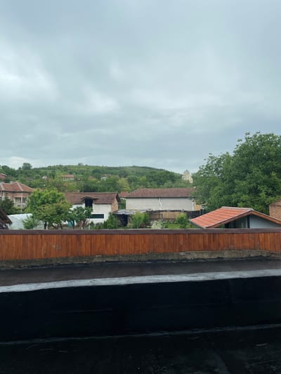 The image shows an overcast view of Rakevo, Bulgaria, from a high vantage point, likely a window or balcony. In the distance, a cluster of buildings and houses with red-tiled roofs are nestled amongst lush green trees and a sloping hillside. On the top of the hill, a yellow dome can be seen. In the foreground is a wooden railing or wall with a portion of roof overhead, which suggests the position is from a building. The sky is uniformly gray, indicating cloud cover. The general atmosphere is quiet and residential.