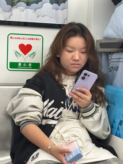 The image depicts a young woman seated inside a public transportation vehicle, possibly a train or subway, in Tiaodeng, China. She is holding a purple smartphone in her right hand and appears to be looking at it intently. A pink and blue portable charger is visible in her left hand, connected to the phone by a light-colored cable. The woman is wearing a white t-shirt, a black and white jacket with ribbed cuffs and collar, and grey pants. She has long, brown, wavy hair. A sign with Chinese characters and an English translation "Courtesy seat" is visible on the wall behind her, indicating a designated seating area for those in need. The interior of the vehicle appears functional and utilitarian, with grey seating and white walls. Other passengers are partially visible in the background.