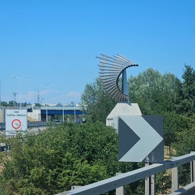 The image shows a contemporary sculpture located in Rozzano, Italy, likely near a highway or major road. The sculpture is made of metal pipes arranged in a fanned-out, semi-circular pattern atop a concrete pedestal. A road sign indicating "zona traffico limitato" (limited traffic zone) with a red circle is visible to the left of the sculpture. Also present is a large, grey and black chevron road sign pointing to the right. In the background, there are trees, overhead road infrastructure including a blue bridge, streetlights, and power lines. The sky is clear and blue. The overall scene depicts an urban or semi-urban environment with elements of transportation and public art.