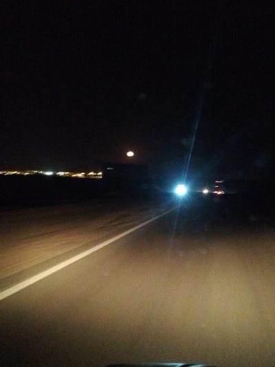 The image shows a nighttime view of a highway in Safaga, Egypt. A white line marks the edge of the lane on the right side of the road. The road appears to be illuminated by the headlights of a car in the foreground, with oncoming headlights in the distance. The lights of the city of Safaga are visible along the horizon in the distance on the left side of the image. Above the lights of Safaga there is a bright, round light that appears to be the moon.