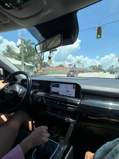 The image shows the interior of a car, looking out through the windshield at a street in Dania Beach, Florida, USA. The driver's hands are visible on the steering wheel and gear shift. A GPS navigation screen is displayed on the dashboard. Outside, palm trees and buildings line the street under a bright, partly cloudy sky. A traffic light is visible, showing a green signal. Several other cars are on the road. The scene suggests a typical daytime drive in a suburban or commercial area.