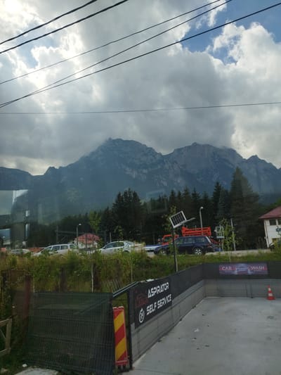 The image captures a view from a window or vehicle, looking out towards a mountainous landscape with a cloudy sky overhead. Power lines crisscross the upper portion of the frame. In the mid-ground, a line of trees separates the foreground from the mountains. Below the trees, several cars are visible, parked or driving along a road. There are also some buildings and a structure that appears to be a self-service car wash. The car wash has signage indicating "ASPIRATOR SELF SERVICE" and "CAR WASH FULL SERVICE." A bright orange traffic cone is placed near the car wash area. The overall scene suggests an urban or semi-urban environment with a backdrop of natural scenery. The location is identified as Bușteni, Romania.