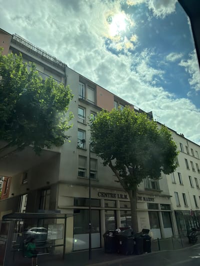 The image shows a building with trees in front of it. The building appears to be an apartment complex or office building with multiple floors and windows. There is a street in front of the building, and some luggage or bags are placed on the sidewalk. The sky is visible in the background, with clouds and the sun shining through.
The building has a sign that reads "CENTRE I.R.M. DE MARNE". The architecture suggests an urban or suburban setting. The presence of luggage might indicate travel or moving. The trees are leafy and green, suggesting it is daytime and likely spring or summer. The sky appears bright, indicating it's daytime, possibly late morning or early afternoon given the sun's position.
The scene is captured from a low angle, looking up at the building and the sky. The overall mood is neutral, presenting a typical street view of a building in Alfortville, France. No people are clearly visible, though the presence of luggage suggests activity nearby. The image is in color and appears to be taken with a camera or smartphone.