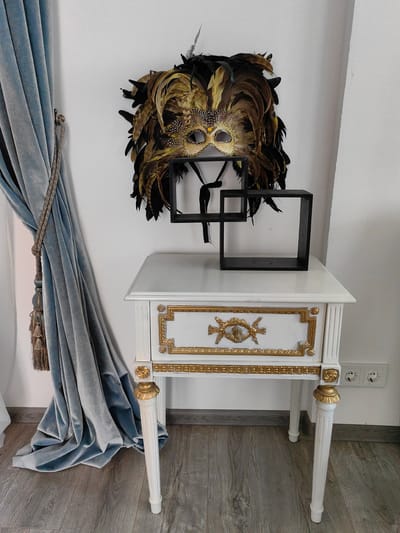 This image captures an indoor scene, likely a room in a home, possibly in Russia given the outlet design. The focus is on a decorative, antique-style white nightstand with ornate gold detailing. Resting on top of the nightstand are two nested, dark-colored geometric display frames, and behind them, a large, elaborate feathered mask is mounted on a white wall. To the left of the nightstand, a luxurious, light blue velvet curtain is drawn back, held by a decorative tassel tieback. The floor is made of wooden planks with a greyish tone. The overall impression is one of elegant, slightly theatrical decor. There is no activity taking place, and the scene is still. The lighting appears to be artificial, suggesting it is either daytime with little natural light or nighttime. No visible text is present.