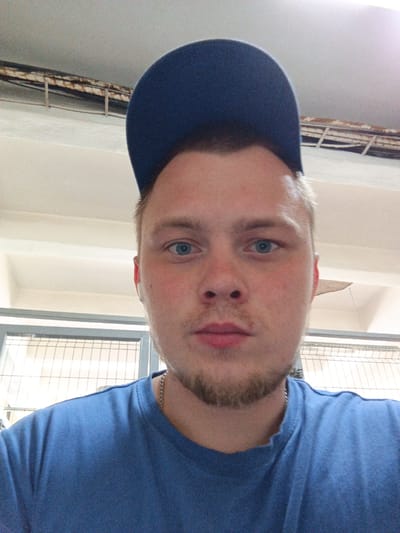 The image is a selfie taken by a young man wearing a blue t-shirt and a blue baseball cap. He has blue eyes and light brown hair and a goatee. The background appears to be indoors, with a white ceiling and exposed pipes or conduit running along the top. There is also a metal fence or railing visible in the lower part of the image. The lighting suggests it might be daytime, possibly with artificial light supplementing natural light. The person's face is the primary focus of the image. No specific location in Shirkovo, Russia, can be identified from the provided image.