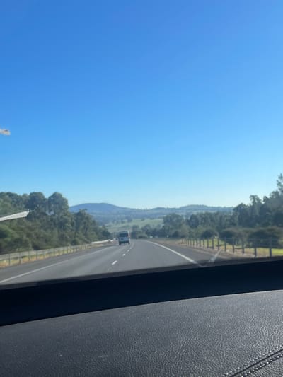 The image depicts a view from the interior of a vehicle traveling on a highway. The foreground is dominated by the dark, textured dashboard of the car. Beyond the dashboard, a multi-lane highway stretches into the distance under a clear, bright blue sky. Trees and rolling hills form the landscape on either side of the road. In the middle ground, a white van is visible on the highway, traveling in the same direction as the vehicle from which the photo is taken. A wooden fence runs along the right side of the highway. The scene suggests a rural or semi-rural environment outside of Taradale, Australia.