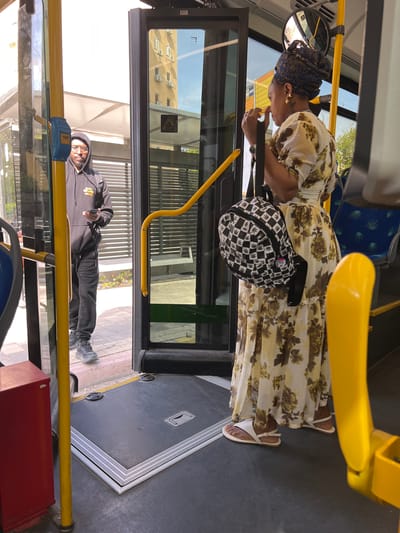 The image captures a scene inside and immediately outside a public bus during what appears to be a bright, sunny day. The bus door is open, suggesting it is either stopped at a station or preparing for passengers to board or alight.
Inside the bus, a woman stands near the open door, facing away from the camera. She wears a long, light-colored dress with a prominent brown and green floral pattern. A dark blue patterned head covering, possibly a scarf or bandana, adorns her head, and she has gold-colored earrings. On her left wrist, a watch is visible. She carries a distinctive black and white checkerboard-patterned backpack, which appears to feature cannabis leaf motifs and a red, white, and blue button or pin. Her footwear consists of light-colored sandals. Her hands are raised near her face, suggesting she might be looking at something small or adjusting something. The bus interior features yellow grab rails, blue patterned seats in the background, and a dark floor with anti-slip strips.
Outside the bus, a man stands on the paved area, looking towards the bus interior. He is dressed in a dark hooded sweatshirt with a partially visible yellow graphic that reads "STAR WARS," dark trousers, and dark sneakers. He wears glasses and holds a small white object in his right hand. His expression is neutral.
Through the bus windows, a building with numerous air conditioning units can be seen in the background, reflecting the clear sky. Visible on the bus window near the man is a hazard symbol (exclamation mark in a triangle) and a word in Hebrew, "שערי" (Sha'arei). The overall setting appears to be an urban or suburban environment.