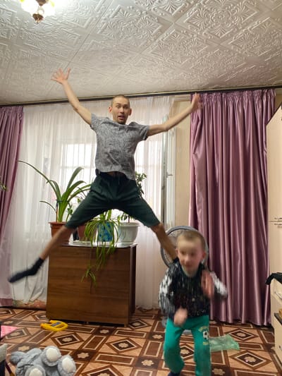 A medium shot, indoor photograph depicts a man and a child in motion within a domestic setting. The man is in mid-air, limbs outstretched as if leaping, positioned in the center. The child, also in motion, appears towards the right side of the frame, seemingly rotating. The room contains a dresser, houseplants, curtains, a fan, toys, and a patterned floor. The photograph's orientation is rotated to the side, as if the camera was held sideways. The location, if in Bely Klyuch, Russia, is inferred from the context of the prompt.