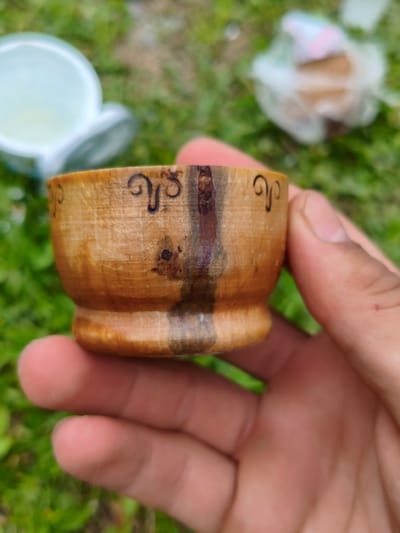 A close-up, outdoor, eye-level shot focuses on a person's hand holding a small, carved wooden cup. The cup is a light brown, with a darker, vertical stripe running down its side, and is decorated with what appear to be carved swirling patterns. The hand holding the cup is that of a light-skinned individual. In the blurred background, green grass is visible, along with out-of-focus elements including a white object with a light blue interior on the left, and a plastic-wrapped object on the right. The image is set in Gusyra, Russia.