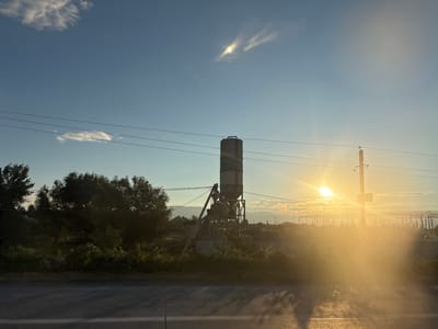 This is a landscape photograph taken in Mukhrani, Georgia. The image depicts a concrete mixing plant silhouette against a bright, hazy sunset. Power lines crisscross the frame, and a utility pole stands to the right. Lush green vegetation and trees fill the middle ground, with a road in the foreground. The sky is a gradient of blue and orange, with wispy clouds and sun rays creating a luminous effect. The overall scene captures an industrial landscape bathed in the warm glow of the setting sun.