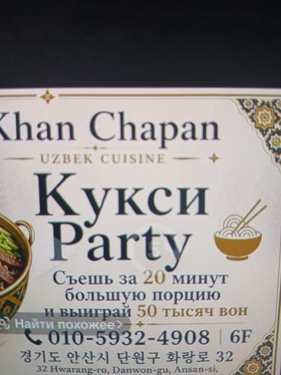 The image is a close-up of a flyer or advertisement for a restaurant named "Khan Chapan Uzbek Cuisine". The flyer is predominantly cream-colored with decorative borders. The main text is in large, dark lettering. The upper portion displays "Khan Chapan Uzbek Cuisine". Below this, in a much larger font, are the words "Кукси Party" written in Russian. Further down, there is Russian text that translates to "Eat for 20 minutes, a large portion, and win 50 thousand won". Below this promotional text, a phone number is displayed: "010-5932-4908 | 6F". Underneath the phone number, Korean text reads "경기도 안산시 단원구 화랑로 32", which translates to "32 Hwarang-ro, Danwon-gu, Ansan-si, Gyeonggi-do". There is a drawing of a bowl of noodles with chopsticks on the right side of the flyer. On the left side, a partially visible illustration of a dish in a woven basket is present. The background of the image is dark, suggesting the flyer is being held or displayed in a dimly lit area. There are no people or specific environmental features of Siheung-si visible in the image itself.