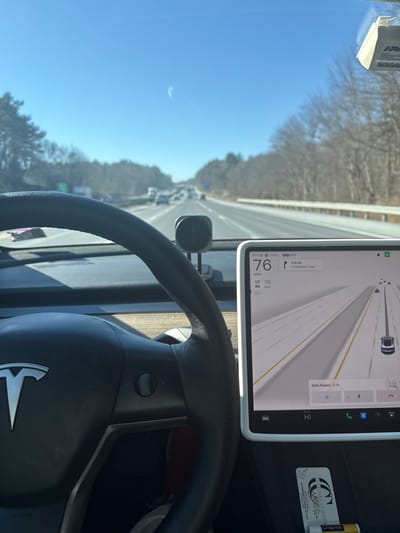 The image captures a first-person perspective from inside a Tesla car driving on a highway during a clear, sunny day. The Tesla steering wheel with its distinctive logo is prominent in the foreground. A large central display unit provides real-time driving information and navigation. The car's speed is indicated as 76 MPH, exceeding the displayed 65 MAX speed limit. The battery level is shown as 76%. Navigation details include "Belle Regina 12:19" as a destination and an upcoming turn onto an "Unnamed road" in 0.8 miles. The display also shows a visual representation of the car's current position on a multi-lane road, surrounded by other vehicles.
Through the windshield, a bright blue sky dominates the upper left, and a highway stretches into the distance, lined with numerous bare deciduous trees, suggesting a winter or early spring season. An EZPass toll transponder is attached to the windshield. On the dashboard, a "Chap-ice" lip balm and a sticker or business card for "COUTURE CORNER MALL" are visible. A phone mount is also present near the base of the central display. The scene depicts active driving on a highway in Charlton, United States.