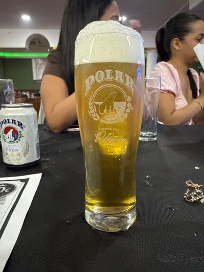 The image shows a close-up of a table at a restaurant in Torrejón de Ardoz, Spain. The focus is on a tall glass of beer with a thick foam head. The beer is amber in color, and condensation is visible on the outside of the glass. To the left of the beer glass, a can of "Polar Pilsen" beer is visible. The can is white with blue and red branding, featuring an image of a polar bear. In the background, two women are seated at the table. The woman on the left has long dark hair and is wearing a dark-colored shirt. The woman on the right is wearing a pink top and has her hair tied back. The table is covered with a black tablecloth. In the lower left corner, a portion of a menu or placemat is visible, with "30€" printed on it. A pair of earrings with rhinestones is also visible on the tablecloth to the right of the beer glass. The restaurant environment appears casual, with a green neon light visible in the background.