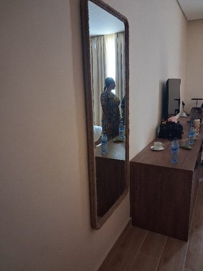 This image depicts an interior room in the city of Takum, Nigeria. A vertical, full-length mirror, featuring a prominent woven or rope-textured frame, is mounted on a plain light beige wall on the left. Reflected in the mirror is the profile of an adult female, facing to the right, dressed in a multicolored patterned garment. Behind her reflection, light-colored curtains are visible, suggesting a window. To the right of the mirror, a dark brown wooden dresser or console table extends horizontally. Arranged on the dresser are three clear plastic water bottles with blue caps, a white ceramic coffee cup with a matching saucer, and a dark-colored bag. Further to the right on the dresser, a dark rectangular electronic device, consistent with a television or monitor, is present. The floor of the room is covered with dark brown tiles featuring a wood-grain pattern.