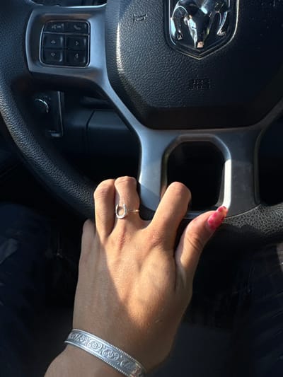 The image depicts a close-up, first-person view from inside a vehicle, focusing on the steering wheel and a human left hand. The steering wheel is black with silver-colored spokes, featuring a prominent chrome Ram truck logo in the center and "SRS AIRBAG" embossed below it. Control buttons for audio and phone functions are visible on the left spoke. A medium-toned human left hand rests on the lower right spoke of the steering wheel. The hand wears a silver-colored horseshoe-shaped ring on the ring finger and a wide, silver-colored bangle with an ornate patterned design on the wrist. The visible thumb has a red/pink, possibly marbled, fingernail. Strong sunlight illuminates the hand and a portion of the steering wheel, casting shadows. The specific location within Rancho Cordova, United States, is not discernible from the visual information provided.