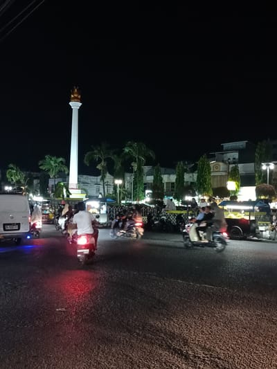 This image depicts a nighttime scene in Bandarbireuen, Indonesia, centered around a large white monument topped with a golden flame-like structure. The monument stands tall against a dark sky, illuminated by artificial lights. Surrounding the monument, the area is bustling with activity, suggesting a public gathering or a lively marketplace.
Several people are visible, primarily on motorcycles, navigating the paved road. Some riders are alone, while others have passengers. The motorcycles' headlights and taillights create streaks of light, adding to the dynamic feel of the image. A white van with a license plate is parked on the left side of the road.
Along the sides of the street, various stalls or small shops are set up, illuminated by lights, further indicating a commercial or social hub. People are gathered around these stalls, some seated, others standing, engaged in conversation or browsing. Palm trees and other foliage are interspersed within the scene, providing a tropical context. The overall atmosphere appears to be one of vibrant nighttime activity, possibly a local gathering or a popular evening spot.
One visible license plate on the white van reads "BL 6332 ZO". The lighting conditions and the presence of multiple illuminated stalls suggest it is nighttime, likely after dusk, with no strong indications of weather conditions. The scene conveys a sense of community and lively social interaction.