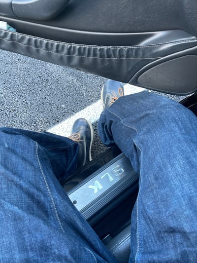 The image displays a downward, first-person perspective from inside a vehicle, focused on the lower body of an individual with an open car door. The subject's legs are covered in dark blue denim jeans. Both feet are shod in dark blue leather boots with light brown laces and visible light-colored soles; the left foot is placed on the ground outside the vehicle, while the right foot remains inside.

The vehicle's interior features a black door panel, which includes a textured pocket or armrest with light stitching, and a speaker grille in the lower section. A metallic door sill plate, prominently displaying "SLK" in silver letters, is situated on the threshold, identifying the vehicle as a Mercedes-Benz SLK model.

The exterior ground surface is dark, coarse-grained asphalt or tarmac. A distinct white painted line runs horizontally across the ground. Direct sunlight casts a sharp shadow diagonally across the asphalt and the subject's footwear. The image lacks identifiable visual markers to confirm the specified location of Plaisir, France.