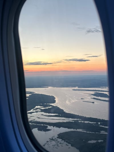 The image captures a view from an airplane window at what appears to be either sunrise or sunset. The sky transitions from a pale blue on the left to warm oranges and yellows as it moves towards the center, suggesting the sun is low on the horizon. Darker, wispy clouds are visible against the lighter sky.
The scene outside the window is a landscape seen from a high altitude. On the right side, a body of water, likely a river or lake, is prominent. Its surface reflects the sky, creating streaks of light and dark. The landmasses bordering the water are dark and appear to be covered in vegetation, possibly forests or trees, given the elongated, textured shapes. The overall impression of the landscape is rugged and natural.
The setting is clearly an airplane, indicated by the dark blue frame of the window in the foreground. The image is oriented vertically, meaning the view is seen from a side window, and the horizon is a vertical line running through the image. There are no people or specific objects clearly identifiable in the landscape, and no text is visible in the image. The time of day is indicated by the sky's colors, and the weather appears clear, with only a few clouds. The location context suggests this view is over Kokkorevo, Russia.