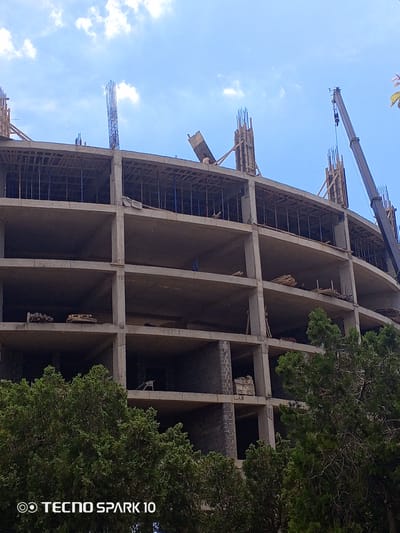 This media file captures a construction scene under a clear blue sky with scattered white clouds. The primary subject is a multi-story building in an unfinished state, characterized by concrete floors, columns, and exposed rebar. Wooden scaffolding and formwork are visible, indicating ongoing construction work. In the background, a large crane with a hook is positioned, suggesting the lifting and movement of materials.

The scene depicts an active construction site, likely during daylight hours given the bright sky and shadows. The presence of trees in the foreground hints at an outdoor environment. While no people are clearly discernible as distinct individuals, there are faint suggestions of human presence within the structure, possibly workers.

Notable details include the curvilinear architecture of the building and the methodical arrangement of construction elements. The weather appears fair, typical for a pleasant day. The location context is Vityazevo, Russia, though no specific landmarks are identifiable in the image itself to confirm this.

The bottom left corner of the image displays text overlay: "TECNO SPARK 10", likely a watermark from the device used to capture the photograph. The overall impression is one of progress and development.