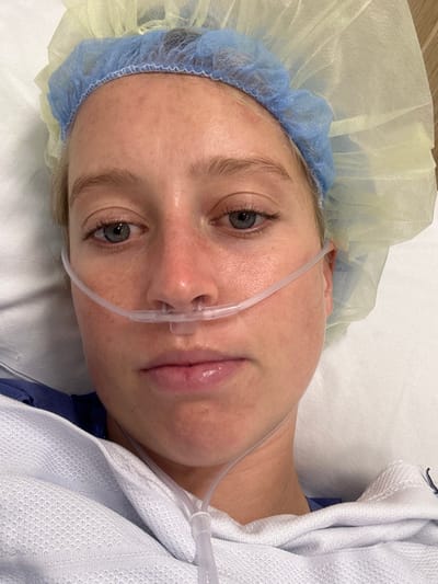A woman with fair skin lies in what appears to be a hospital bed. She wears a blue and yellow surgical cap and a nasal cannula delivering oxygen. Her eyes are open, and her gaze is directed toward the camera. A white blanket is draped over her shoulder. The woman's face shows some redness, suggesting recent medical procedures. Her lips appear slightly swollen. The overall impression is one of post-operative recovery.