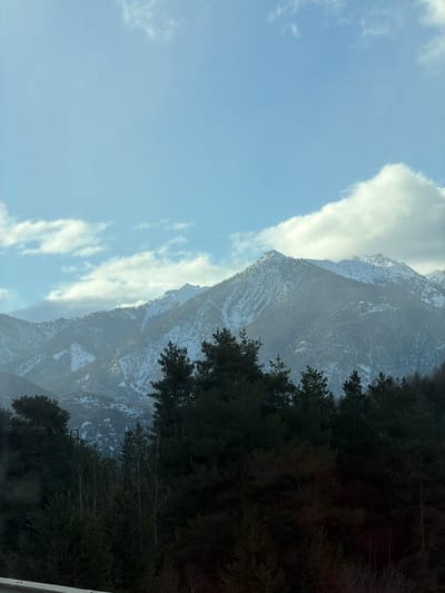 The image depicts a snow-dusted mountain range adjacent to a forest. A light blue sky with scattered clouds is visible on the left. A row of trees is present on the right, partially obscuring the view of the mountains. The photo appears to be taken from a lower elevation, possibly from a road. The location is Saint-Chaffrey, France.