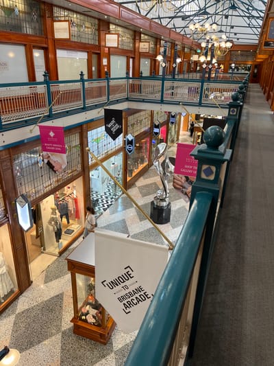 The image displays an elevated view inside a grand, multi-level indoor shopping arcade, characterized by its ornate heritage architecture. The perspective is from an upper floor, looking down the length of the arcade, which features rich wooden paneling on storefronts with large glass display windows on both levels. Decorative white railings with teal-green posts and accents line the walkways. Overhead, multiple-bulb chandeliers illuminate the space, suggesting it is daytime, possibly complemented by natural light from a skylight.

In the center of the lower level, a large, silver, abstract sculpture resembling the "Spirit of Ecstasy" sits on a black pedestal, serving as a prominent display. The floor below is tiled in a distinctive black and white checkerboard pattern. Various banners hang throughout the arcade: a pink banner advertises "BEAUTY SERVICES GALLERY LEVEL" and another, partially visible, reads "SHOP THE GALLERY LE". A black banner mentions "MADE IN BRISBANE ARCADE," while a white banner prominently declares "UNIQUE TO BRISBANE ARCADE."

Shop signs identify businesses such as "KIMMISON JEWELLERY" and "Glamor Beauty Services," along with a partially visible "CAMERON SILVER" sign. At least one person can be seen on the lower level, near a shop entrance, adding to the sense of retail activity. The scene depicts a calm and elegant shopping environment in Brisbane City, Australia, specifically within the Brisbane Arcade.
