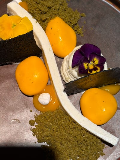 The image depicts a plated dessert presentation. The dessert elements include several yellow, spherical components resembling mango or fruit gelées; a rectangular white mousse or meringue bar; a dark, rectangular, possibly savory component; a light green crumble; a swirl of white cream, and two edible flowers (one yellow, one purple). The dish is presented on a dark, possibly ceramic or stone plate. No people are visible. The location in Kiryat Ono, Israel is not evident from the image itself.