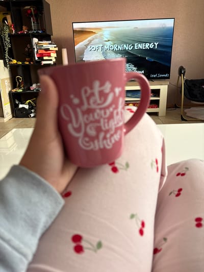 A person is holding a pink mug with white cursive writing in front of a pink cherry-patterned pillow. The mug is slightly out of focus. Behind the person and mug is a room with a wall-mounted TV showing a nature scene, a white shelf, and a black shelving unit with books and other objects. The setting appears to be indoors. There is no information provided in the image that indicates this location to be Veghel, Netherlands, nor anything that could suggest this.