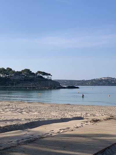 The image depicts a tranquil beach scene in Peguera, Spain, under a clear blue sky. The foreground is dominated by a sandy beach with tire tracks from a vehicle, likely a beach groomer, and some footprints. A wooden boardwalk extends into the frame from the lower right. The middle ground features calm, clear turquoise water, with several yellow buoys marking a designated swimming area. A single person is visible wading in the water. In the background, a rocky outcrop covered with pine trees and a cluster of white buildings, possibly hotels or apartments, is situated on a hill overlooking the sea. Further in the distance, a coastal town with a dense arrangement of buildings climbs up a hill, and a prominent tower or structure is visible on its summit. The overall impression is one of a peaceful and idyllic coastal resort.