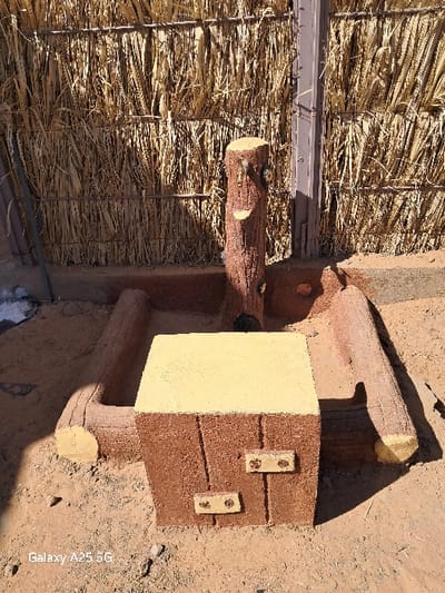 The image shows a small, rustic, outdoor structure, possibly a well or water pump, made of stacked logs and cement. It is located in a courtyard or backyard setting, situated against a wall constructed of woven straw mats. The wall is made of brown, dried grass, interwoven to form a fence-like barrier. The structure features a square platform with a yellow top and two wooden-looking, brown-painted doors. A single wooden post, resembling a weathered tree trunk, stands upright in the center of the structure. The scene is bathed in sunlight, casting shadows on the ground and the structure. The environment is dry and dusty, suggesting a desert location. The ground is primarily sandy, with patches of dirt.  The location, based on the coordinates provided, is in Karima, Sudan. There are no people in the scene.