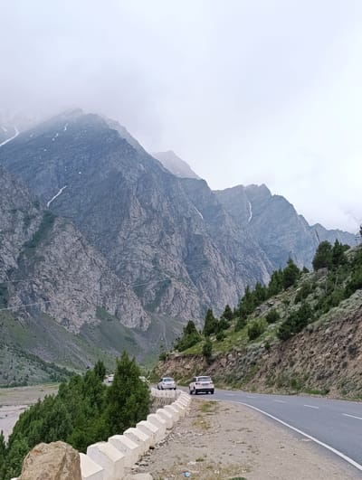 The image depicts a scenic mountain road in Darcha, India. A winding asphalt road with a white line marking the lanes curves through a mountainous landscape. On the right side of the road, a steep, rocky slope is covered with green vegetation and scattered trees. A low, white concrete barrier with a scalloped design separates the road from the embankment.
Two white cars are visible on the road. One car is parked or moving slowly on the right side, partially obscured by the barrier. Another car is further ahead on the left side of the road, also white. In the background, imposing, rugged mountains rise steeply. These mountains are characterized by their rocky, textured surfaces and are partially shrouded in clouds at their peaks, with patches of snow visible on their slopes. The sky is overcast and hazy, creating a muted, diffused light. The valley floor to the left of the road appears to be a dry riverbed or plain with sparse vegetation and a few trees. The overall scene conveys a sense of remote, natural beauty in a high-altitude environment.