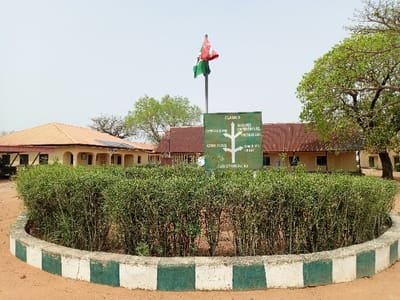 The image shows a courtyard at the Federal College of Education (Technical) in Rumde Waziri, Nigeria. It is a sunny day with a clear sky. The courtyard is surrounded by a low wall and a circle of green bushes. A signpost at the center of the circle indicates directions to various facilities. The sign reads: "CLASSER", with arrows pointing towards "COMPUTER CENTRE", "ADMIN BLOCK", "OUTLICK CHEMISTRY LAB", "BOYS HOSTEL", "GIRLS HOSTEL", and "GARBAGE SCENES". A Nigerian flag is waving on a pole to the left of the sign. There are a few buildings with brown tile roofs visible behind the bushes. The scene is a typical courtyard within a campus environment.