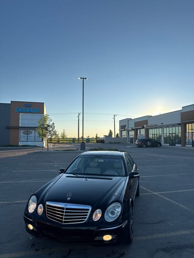In Edmonton, Canada, at approximately 6:30 PM MDT, a black Mercedes-Benz E-Class sedan is parked in a paved parking lot. The car is positioned at an angle, facing slightly towards the viewer, with its front end and driver's side visible. The vehicle has its headlights and fog lights illuminated.

Behind the car, a row of commercial buildings lines the street, featuring large glass windows and signage. Notable businesses include "Ewing Carwash" on the left and "Putts & Paws" further down the row. The architecture suggests a modern strip mall or commercial complex. A tall, dark lamppost stands near the center of the image, its light on.

The environment is an open-air commercial area, likely a parking lot for a shopping center or business park. The sky is a clear, pale blue, transitioning to a lighter hue near the horizon where the sun is setting, casting a warm glow. A few trees are scattered throughout the scene, and power lines are visible strung between utility poles. The asphalt of the parking lot is marked with yellow painted lines indicating parking stalls. The overall scene depicts a common urban commercial landscape during twilight.