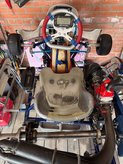 The image shows a go-kart parked indoors, likely in a garage or workshop, with a brick wall and wooden flooring visible in the background. The go-kart is viewed from above and is the central subject. It is white and blue with red and blue accents on the steering wheel. The steering wheel has a digital display. The seat is white and appears worn. The go-kart has a Rotax engine, visible on the right side of the image, which is red and black. The rear wheels and exhaust system are also visible. The overall scene is static, with no people present or any apparent activity. The environment suggests a utilitarian space, possibly associated with racing or go-kart maintenance. The image does not contain any visual cues that would indicate its location in Sukhaya, Russia.