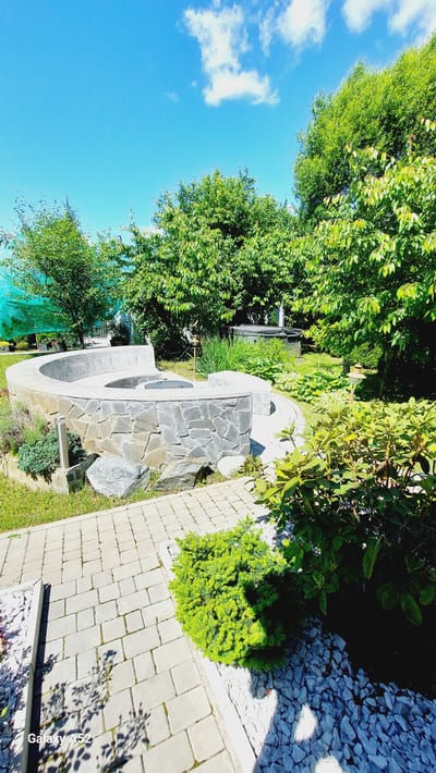 The image depicts a circular stone bench with a fire pit in the center, situated in a garden in the city of Zhavoronki, Russia. The bench is constructed from large, irregular grey stones. Behind the bench, lush green trees and various shrubs create a natural backdrop. The ground is paved with light grey interlocking stones, and there are patches of grass and decorative grey gravel. A bright blue sky with scattered white clouds is visible above. In the background, a hot tub or spa can be seen amongst the foliage. The scene suggests a well-maintained outdoor recreational area.